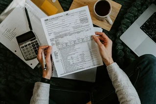 An individual holds a 1040 Tax Form with various documents surrounding them, a calculator, a laptop and coffee