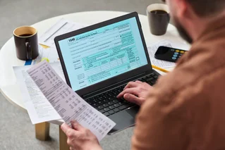 Over the shoulder shot of a man holding a receipt and typing into a tax form on his laptop.