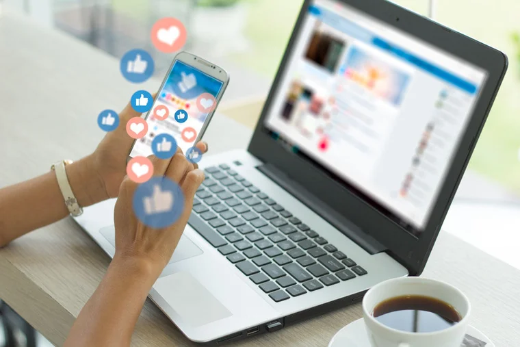 An individual holds their phone in their hand in front of their laptop, social media platforms are visible on each as the "like" and "heart" emoji icons float from the screen