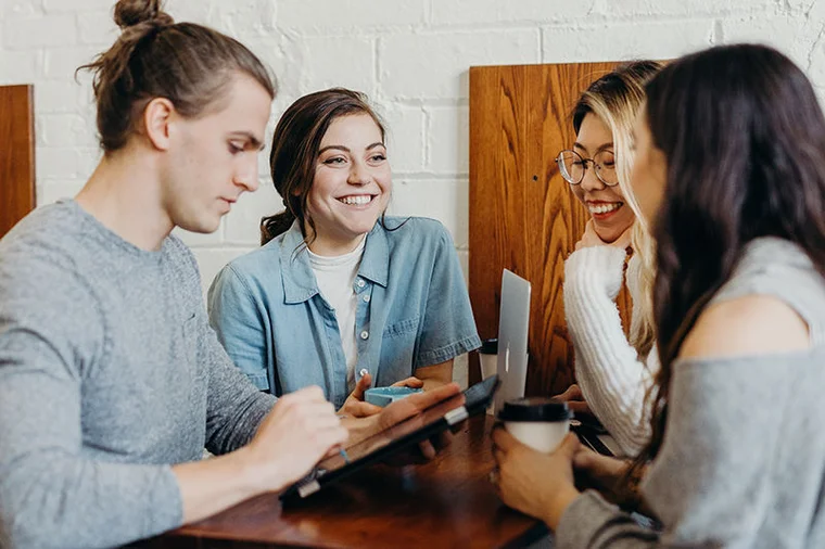 A group of young adult friends smiling and gathering around a tablet and laptop